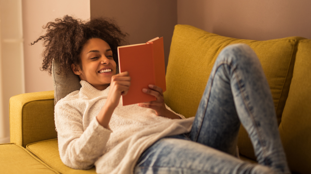 Woman lies on couch happily reading a paper book. Reading is good for children's mental health and adult mental health.