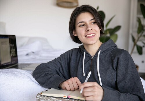 woman holding pen with hands on top of notebook sits next to open laptop